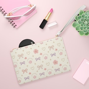 May include: Cream-colored clutch with a repeating pattern of pink and purple bows, hearts, and flowers. The clutch has a gold zipper and a white wrist strap. Other items on the pink surface include sunglasses, lipstick, and a succulent.