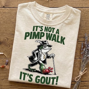 May include: Cream-colored t-shirt with a humorous graphic. The design features a frog in a fedora and coat, walking with a cane, and the text "IT'S NOT A PIMP WALK IT'S GOUT!" in green. The shirt has a Comfort Colors tag.