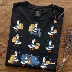 May include: Black t-shirt featuring a whimsical design of cartoon geese delivering packages. The geese are depicted in blue uniforms, carrying boxes, riding a scooter, and driving a delivery van. The shirt has a Comfort Colors tag.