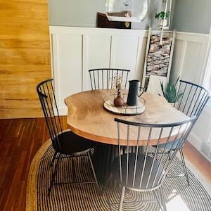 May include: A dining room set featuring a round wooden table with a live edge, surrounded by four black metal chairs. The table is set on a circular rug with black and beige stripes. A clear glass pendant light hangs above the table.