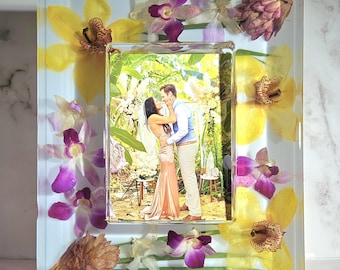 May include: A clear resin photo frame with a picture of a couple kissing. The frame is decorated with yellow and purple orchids, and other dried flowers. The frame is rectangular with a clear border.