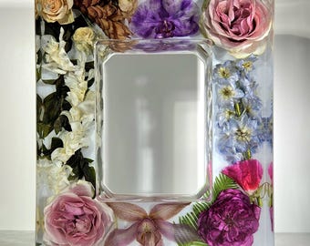May include: A rectangular photo frame with a clear resin border, embedded with a variety of dried flowers in shades of pink, purple, white, and yellow. The frame has a rectangular opening for a photo.