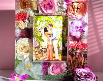 May include: A clear resin photo frame with embedded dried flowers, including roses and orchids. The frame holds a photo of a couple embracing. The frame is rectangular with a smaller rectangular opening for the photo.