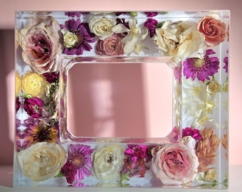 May include: A rectangular clear resin picture frame with dried flowers embedded within. The flowers include roses, daisies, and other blooms in shades of pink, purple, and cream. The frame has a rectangular opening.