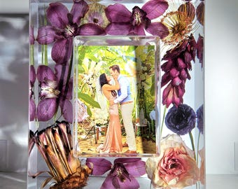 May include: A clear resin photo frame with a rectangular opening, showcasing a couple embracing. The frame is adorned with embedded purple flowers and other dried floral elements, creating a unique decorative piece.
