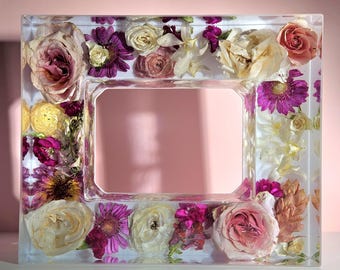 May include: A rectangular clear resin picture frame with dried flowers embedded within. The flowers include roses, daisies, and other blooms in shades of pink, purple, and cream. The frame has a rectangular opening.