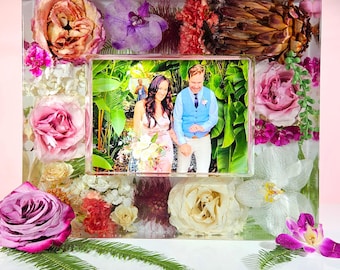 May include: A clear resin photo frame with embedded flowers, including roses and orchids. The frame holds a photo of a couple. The frame is rectangular with a smaller rectangular opening for the photo.