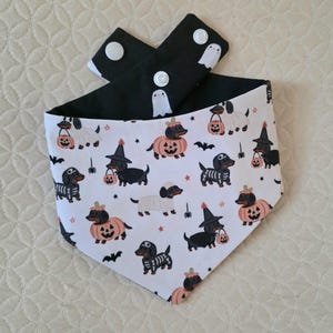 May include: A triangular pet bandana featuring a white background with a repeating pattern of dachshunds in various Halloween costumes, including pumpkins, skeletons, and witch hats. The reverse side is black with white ghost icons and white snaps.