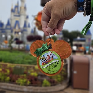 May include: A circular button with a green background featuring an illustration of Goofy and the text "I'm Celebrating: MNSSHP" and "Walt Disney World". The button is set in an orange pumpkin-shaped frame with a glittery green bow attached.