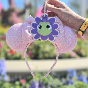 May include: Pink sequined Minnie Mouse ears headband with a purple flower charm featuring a green frog face.