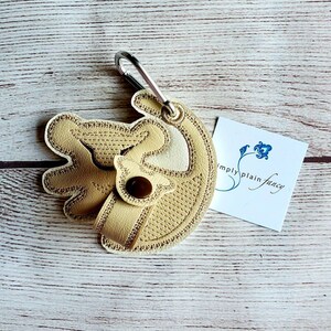 May include: A beige leather keychain with a stitched design of a dog's head. The keychain has a silver carabiner clip and a brown button. The keychain is attached to a white card with the text "simply plain fancy".