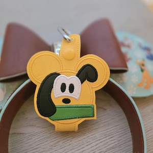 May include: A yellow faux leather keychain featuring an embroidered character face with large rounded ears, a black floppy ear, white eye patches, and a green rectangular accent. The keychain includes a silver metal clasp and a snap button.