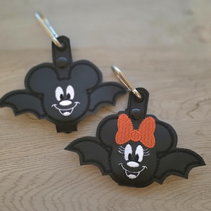 May include: Two black bat-shaped keychains featuring embroidered character faces. One keychain displays a white face with round ears, while the other features a white face with a bright orange bow. Both include a silver metal lobster clasp attachment.