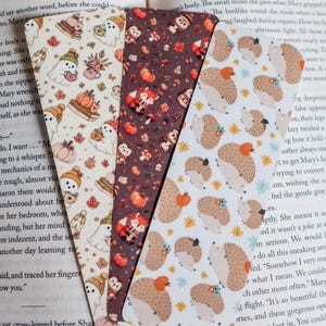 May include: Three rectangular bookmarks featuring autumn-themed patterns. The left bookmark has a cream background with ghosts, pumpkins, and leaves. The center is dark brown with foxes and pumpkins. The right is white with hedgehogs and leaves.