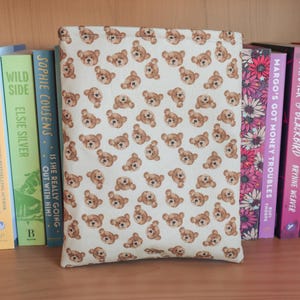 May include: A cream-colored book sleeve featuring a repeating pattern of brown teddy bear faces. The sleeve is positioned among a collection of colorful books on a wooden shelf.