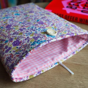 May include: A floral fabric book sleeve with a button closure. The exterior features a pattern of purple, blue, and yellow flowers on a white background. The interior lining is pink gingham. A red book with the text "BEST OFFER WINS" is in the background.