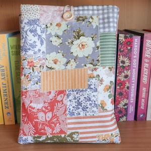 May include: A patchwork book sleeve with floral and striped patterns in shades of red, blue, and cream. The sleeve is secured with a wooden button and loop. Books are visible in the background.