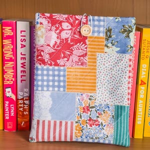 May include: A patchwork fabric book cover with a button closure, resting among a row of colorful books. The cover features a mix of floral, striped, and gingham patterns in red, blue, and orange. The books have titles like "Mr. Wrong Number" and "Nothing Like the Movies."