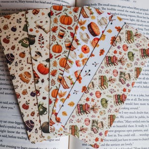 May include: A set of five rectangular bookmarks featuring various autumn-themed patterns including orange pumpkins, colorful fall leaves, stacks of books, warm beverages in mugs, and cozy sweaters printed on a light cream-colored background.