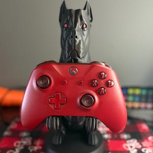 May include: A black dog-shaped controller stand holding a red Xbox controller. The controller has the Xbox logo and the A, B, X, and Y buttons. The dog stand has red eyes.