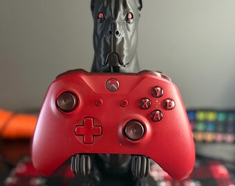 May include: A black dog-shaped controller stand holding a red Xbox controller. The controller has the Xbox logo and the A, B, X, and Y buttons. The dog stand has red eyes.