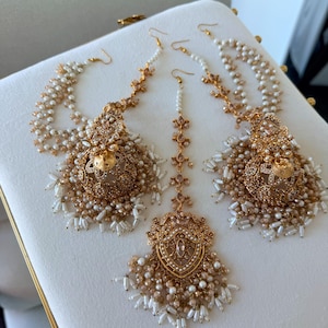 May include: A set of gold-toned jewelry consisting of two large jhumka earrings with intricate filigree, dangling white and champagne-colored beads, and a matching central maang tikka headpiece featuring a teardrop-shaped pendant design.