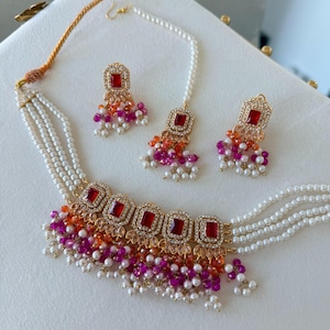 Può includere: Un set di gioielli composto da una collana di perle a più fili, due orecchini abbinati e un maang tikka. Ogni pezzo presenta pietre rosse rettangolari incorniciate da cristalli chiari, con perline pendenti di colore arancione e rosa.