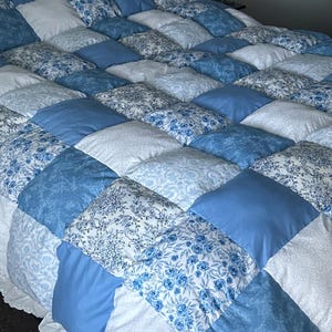 Op de afbeelding: Een handgemaakte patchwork quilt in blauwe en witte tinten. De quilt heeft vierkante blokken met bloempatronen en effen kleuren, afgewerkt met een geschulpte rand. De quilt ligt op een zwart bedframe.