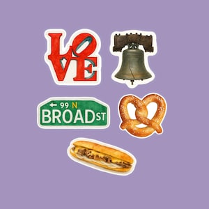 May include: A collection of stickers featuring Philadelphia-themed designs. Includes a red and white "LOVE" sign, the Liberty Bell, a street sign reading "99 N BROAD ST", a pretzel, and a cheesesteak sandwich.
