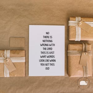 Peut inclure: Une carte blanche avec du texte noir qui dit "NO THERE IS NOTHING WRONG WITH THE CARD THIS IS JUST WHAT WORDS LOOK LIKE WHEN YOU GET THIS OLD" est entour&eacute;e de cadeaux emball&eacute;s et d'une petite carte.