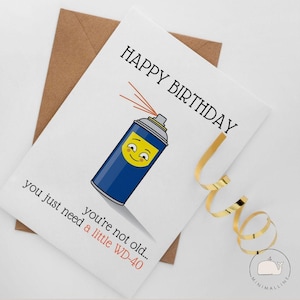May include: A white greeting card featuring a blue spray can illustration with a yellow smiling face. The text reads HAPPY BIRTHDAY and you're not old... you just need a little WD-40. The card is paired with a brown envelope and gold ribbon.