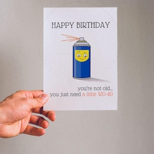 May include: A white greeting card featuring a blue spray can illustration with a yellow smiling face. The text reads HAPPY BIRTHDAY at the top and you're not old... you just need a little WD-40 at the bottom in black and red lettering.