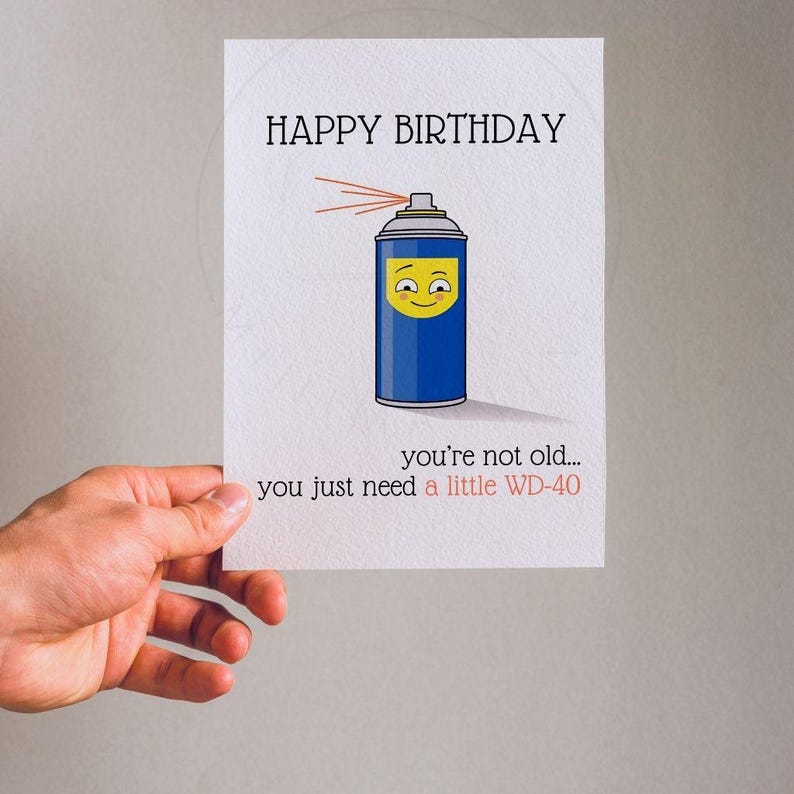 May include: A white greeting card featuring a blue spray can illustration with a yellow smiling face. The text reads HAPPY BIRTHDAY at the top and you're not old... you just need a little WD-40 at the bottom in black and red lettering.