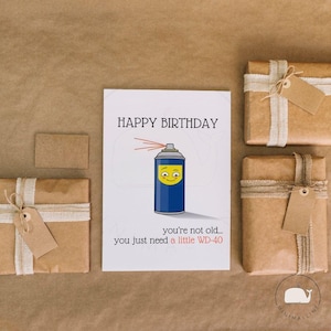 May include: A white rectangular greeting card featuring a blue spray can illustration with a smiling face and the text Happy Birthday you're not old... you just need a little WD-40. The card is surrounded by three brown paper wrapped gift boxes.