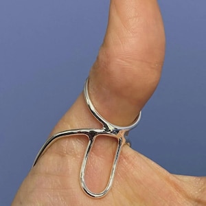 May include: A silver-colored ring with a unique design, featuring a curved band and a central, elongated loop. The ring is worn on a thumb, with a blue background.