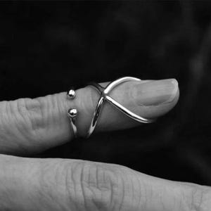 May include: A silver-toned ring with an X-shaped design and a separate band with two small spheres. The ring is worn on a finger, showcasing a modern and minimalist jewelry style. The image is in black and white.