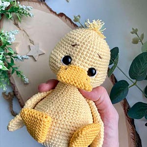 Puede incluir: Un juguete de patito tejido a mano en tonos amarillos. El patito tiene un pico amarillo, ojos negros y pies pequeños. El juguete se sostiene en una mano, con una rodaja de madera y estrellas decorativas en el fondo.