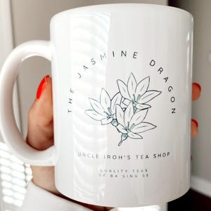 Op de afbeelding: Witte keramische mok met de tekst "THE JASMINE DRAGON" en "UNCLE IROH'S TEA SHOP" in zwart. Een lichtblauwe bloemenillustratie is gecentreerd op de mok. De mok heeft ook de tekst "QUALITY TEAS OF BA SING SE".