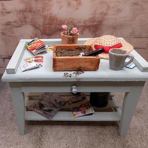 Puede incluir: Una escena de jardinería en miniatura sobre una mesa azul claro. Incluye una jardinera de madera con tierra, una pequeña paleta, un sombrero de paja con una cinta roja, una taza de café pequeña, sobres de semillas y una maceta con flores rosas.