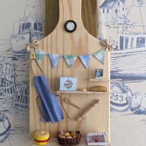 May include: A wooden cutting board with a miniature bakery scene. The board has a clock, a blue and white striped apron, a rolling pin, a wooden spoon, a box of pastries, a basket of bread rolls, and a box of cookies. The board is decorated with a blue and green bunting.
