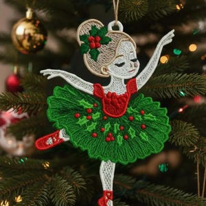 Op de afbeelding: Geborduurde kerstversiering van een ballerina in een groene tutu met rode accenten. De ballerina heeft een blonde knot met een hulst- en bessenaccent. De versiering hangt aan een kerstboom met andere versieringen.