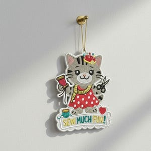 以下が含まれることがあります： 「SEW MUCH FUN!」の文字が入った、猫の形をした刺繍オーナメント。猫は黒い縞模様のグレーで、赤い水玉模様のドレスを着て、糸巻きとハサミを持っています。吊り下げ用の金色の紐が付いています。