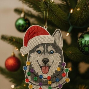 May include: A Christmas tree ornament shaped like a smiling husky dog wearing a Santa hat and a string of colorful lights. The ornament is gray, white, and black with blue eyes and a red tongue.