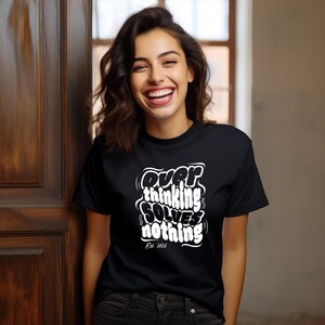 Puede incluir: Camiseta negra con el texto blanco "Overthinking Solves Nothing" y "Est. 2018". La camiseta la lleva una persona con cabello oscuro y una sonrisa.