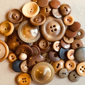 Può includere: Una collezione di bottoni vintage in varie tonalità di marrone e beige. I bottoni sono realizzati con diversi materiali, tra cui plastica, metallo e legno. Alcuni dei bottoni hanno disegni intricati, mentre altri sono semplici. I bottoni sono disposti in uno schema casuale su una superficie marrone chiaro.