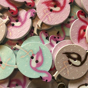 Könnte beinhalten: Eine Sammlung von Holz-Knöpfen mit pinken Flamingos in verschiedenen Posen. Die Knöpfe sind in Rosa-, Blau- und Weißtönen bemalt, einige haben eine glitzernde Oberfläche.