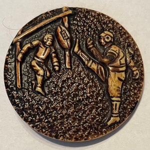 Puede incluir: Un botón de cerámica marrón con una imagen en relieve de dos jugadores de fútbol. Un jugador está pateando un balón de fútbol hacia una portería mientras que el otro jugador corre hacia la portería.