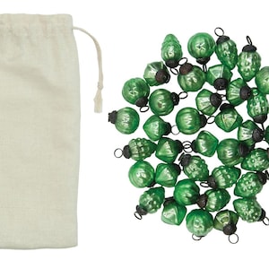 May include: A collection of small, green glass Christmas ornaments in various shapes, with black metal caps. A beige drawstring bag is on the left. The ornaments are scattered on a white background, suggesting a festive holiday theme.