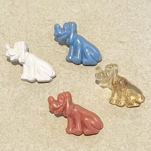 Op de afbeelding: Vier plastic knopen in de vorm van honden in verschillende kleuren: wit, blauw, bruin en transparant. De knopen zijn allemaal in dezelfde pose, met hun hoofd naar de zijkant gekanteld en hun tongen uitgestoken.