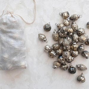 May include: A collection of vintage-style silver ornaments, some scattered and some inside a small, off-white drawstring bag. The ornaments have various shapes and textures, with a metallic finish. The background is a textured, light-colored surface.